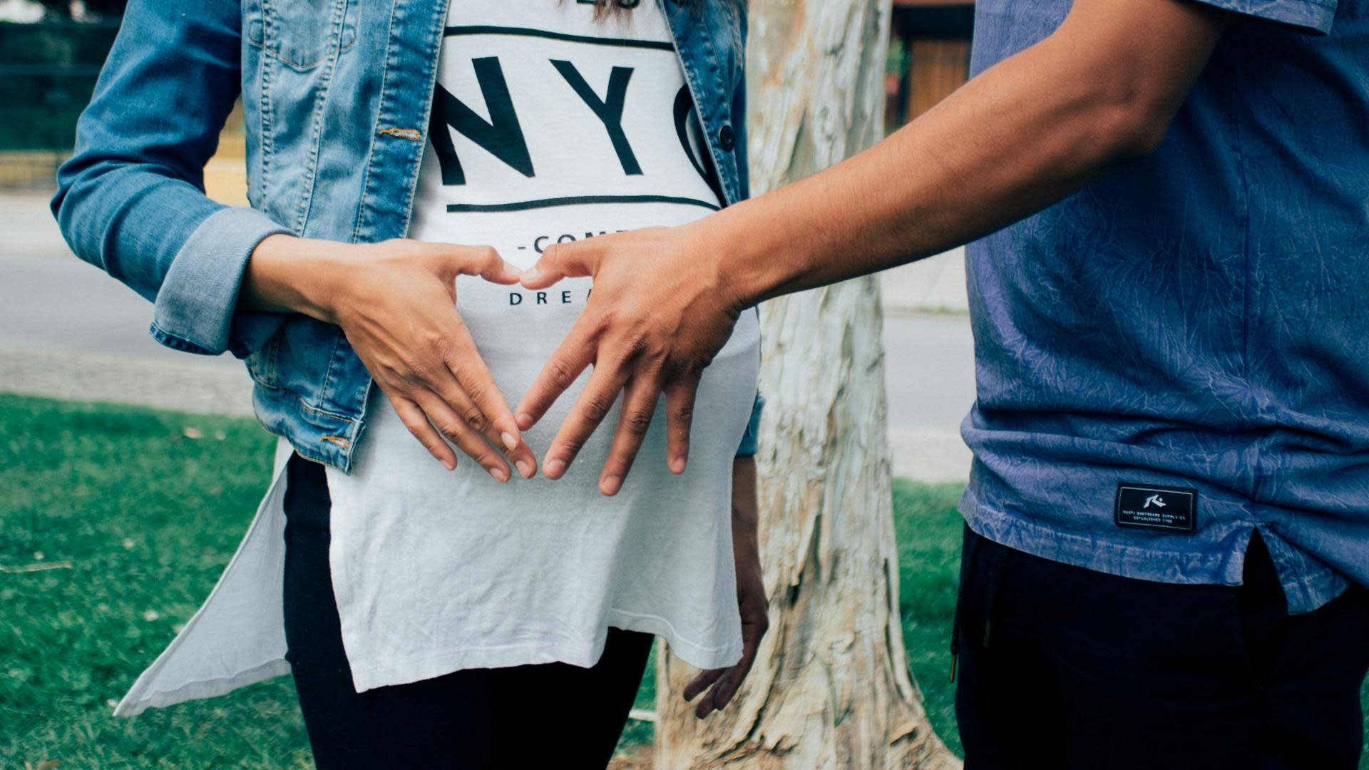 Two people, one in a denim jacket and white shirt, and the other in a blue shirt, stand forming a heart shape with their hands over the first person's baby bump. They are outdoors, next to a tree and grass area, celebrating the joy of pregnancy-related benefits like paid prenatal care leave.