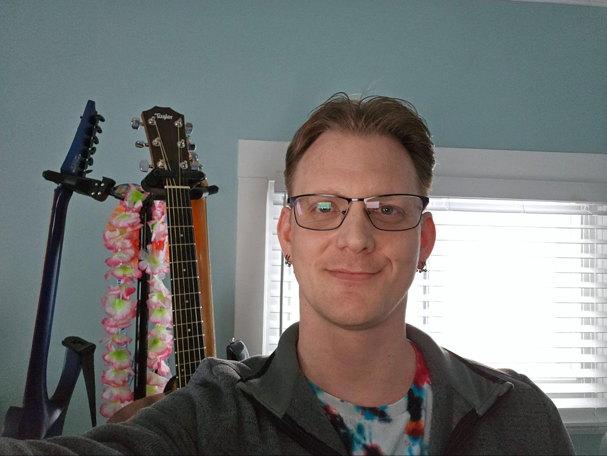 A person with short hair, wearing glasses and a tie-dye shirt, smiles at the camera. Behind them are guitars positioned on stands, with one guitar adorned with a colorful floral lei.